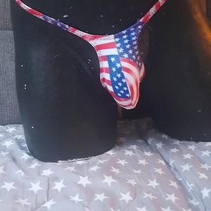 Könnte beinhalten: Ein String-Badeanzug mit dem Design der amerikanischen Flagge. Der Stoff zeigt die roten und weißen Streifen und das blaue Feld mit weißen Sternen. Das Kleidungsstück wird auf einer schwarzen Schaufensterpuppe präsentiert.