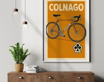Op de afbeelding: Een ingelijste poster met een illustratie van een oranje Colnago-fiets. Het kunstwerk toont een racefiets tegen een oranje achtergrond, met het Colnago-logo en tekst. De poster zit in een witte lijst.