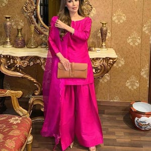May include: A vibrant fuchsia pink ensemble, featuring a long-sleeved tunic and wide-leg pants. A matching scarf and a tan clutch complete the outfit. The clothing is made of a shiny fabric.