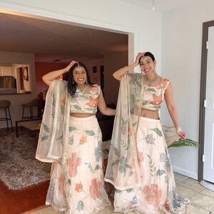 May include: Two women wearing matching cream-colored traditional Indian outfits. The outfits include a crop top, a long skirt, and a sheer shawl. The outfits are decorated with floral patterns in shades of pink, orange, and blue.