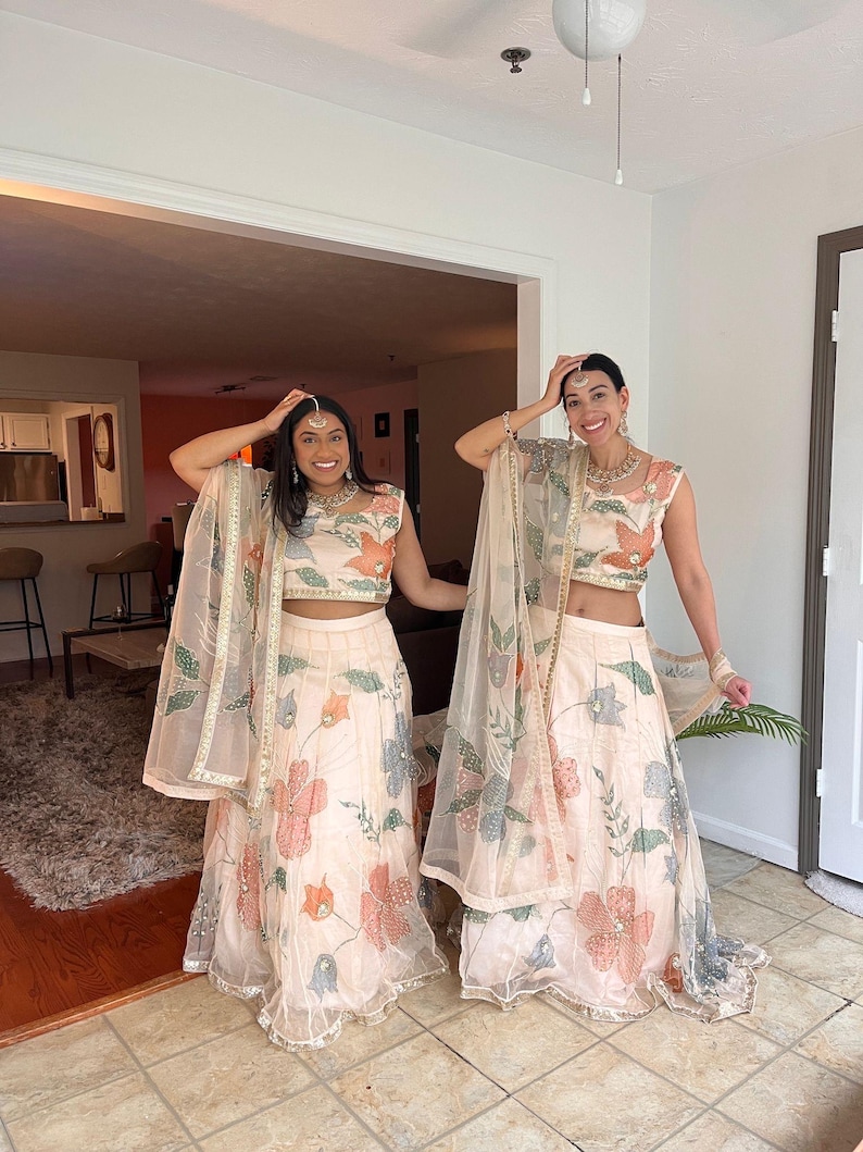 May include: Two women wearing matching cream-colored traditional Indian outfits. The outfits include a crop top, a long skirt, and a sheer shawl. The outfits are decorated with floral patterns in shades of pink, orange, and blue.