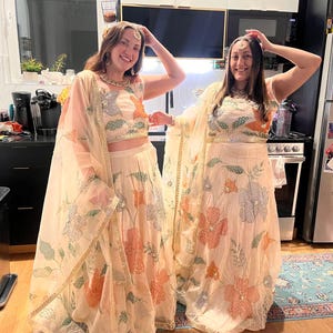 May include: Two people wearing matching cream-colored outfits with floral embroidery. The outfits include crop tops, skirts, and sheer scarves. The floral designs feature orange, blue, and silver accents. They are smiling and posing in a kitchen.