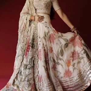 May include: An ivory-colored, floral-printed lehenga with a matching blouse and dupatta. The skirt features large pink and orange flowers with silver accents. The blouse and dupatta have intricate embroidery. The outfit is suitable for special occasions.