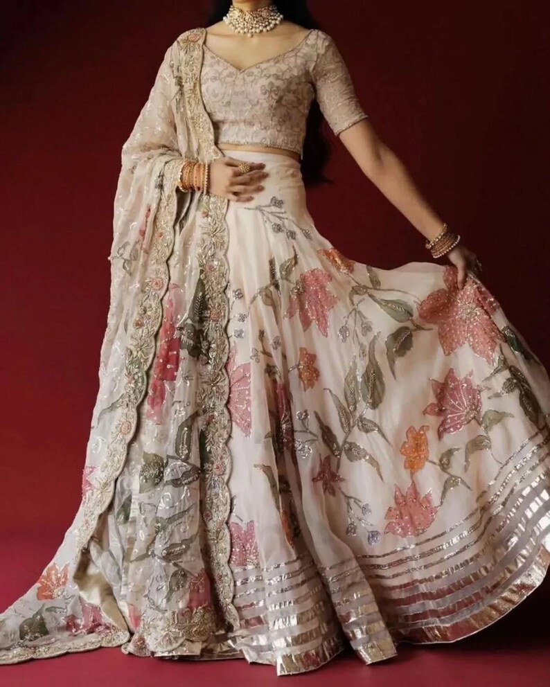 May include: An ivory-colored, floral-printed lehenga with a matching blouse and dupatta. The skirt features large pink and orange flowers with silver accents. The blouse and dupatta have intricate embroidery. The outfit is suitable for special occasions.