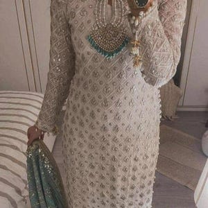 May include: Ivory-coloured, long-sleeved dress with intricate embroidery and embellishments. The dress features a keyhole neckline adorned with pearls and turquoise accents. A matching teal scarf is held in the hand. The dress is a formal wear garment.