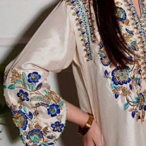 Pu&ograve; includere: Tunica ricamata color crema con motivi floreali blu e oro. Il polsino della manica presenta ricami dettagliati. Un braccialetto dorato adorna il polso. L'indumento ha un design fluido ed elegante.