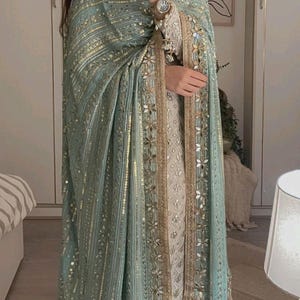 May include: A teal and gold embroidered shawl with detailed embellishments. The shawl has a delicate pattern of sequins, creating a luxurious look. The garment is draped over a cream-colored outfit, showcasing the contrasting colours and textures. The outfit is suitable for special occasions.