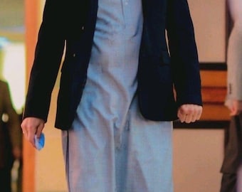 Op de afbeelding: Een persoon draagt een zwarte blazer over een lichtblauwe tuniek en bijpassende broek. De outfit heeft een mandarijnkraag en een knoopsluiting. De persoon houdt een klein, opgerold blauw voorwerp vast.