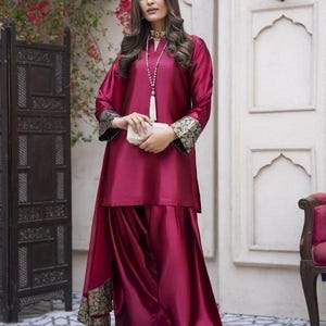 May include: A burgundy satin ensemble featuring a long-sleeved tunic with gold and black embroidered cuffs, paired with matching wide-leg pants. The outfit includes a long pearl necklace and a small white clutch. Gold heels complete the look.