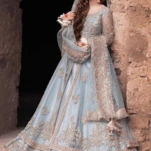 May include: A light blue formal gown with intricate gold embroidery and embellishments. The dress features long sleeves, a flowing skirt, and a matching veil. The gown is suitable for special occasions.