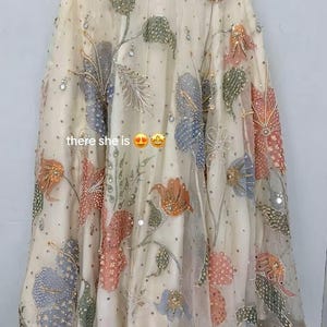 May include: A cream-colored, sheer garment with intricate floral embroidery in shades of orange, pink, blue, and green. The design includes sequins and the text "there she is". The hemline is detailed with gold embellishments.