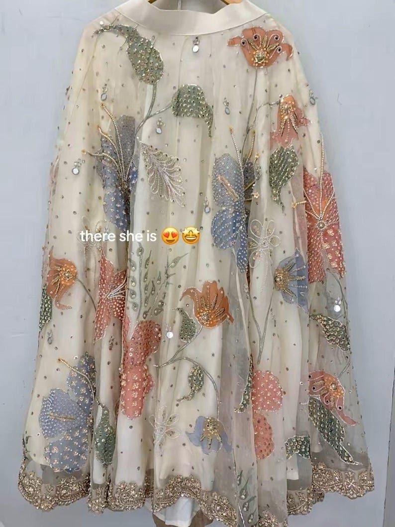 May include: A cream-colored, sheer garment with intricate floral embroidery in shades of orange, pink, blue, and green. The design includes sequins and the text "there she is". The hemline is detailed with gold embellishments.
