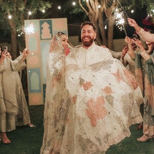 May include: A smiling couple celebrates their wedding, the groom carrying the bride. The bride's white dress features pink floral embroidery and silver embellishments. Guests hold sparklers, creating a festive atmosphere.
