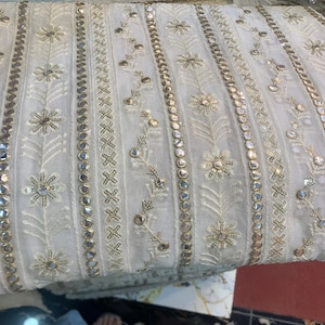 May include: Close-up of white fabric with detailed embroidery and embellishments. The design includes floral and geometric patterns, with gold-coloured accents and small, reflective mirror-like details. The fabric is folded, highlighting its texture and design.