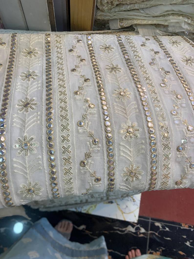 May include: Close-up of white fabric with detailed embroidery and embellishments. The design includes floral and geometric patterns, with gold-coloured accents and small, reflective mirror-like details. The fabric is folded, highlighting its texture and design.