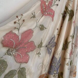 May include: A sheer, cream-colored fabric adorned with embroidered floral designs. Large pink flowers with silver detailing and smaller green leaves are scattered across the material. The fabric is draped, showcasing the intricate embroidery and delicate texture.
