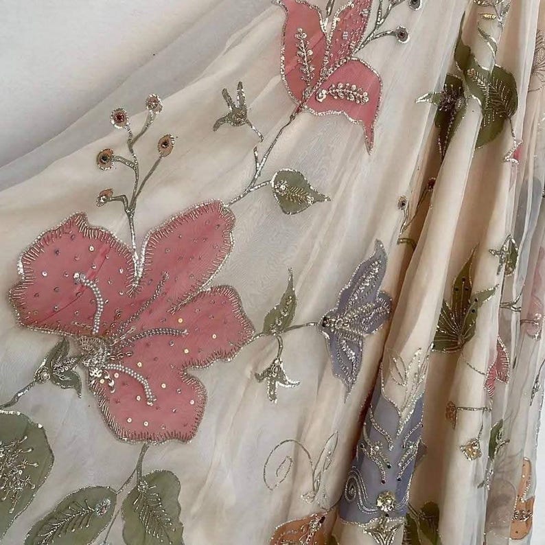 May include: A sheer, cream-colored fabric adorned with embroidered floral designs. Large pink flowers with silver detailing and smaller green leaves are scattered across the material. The fabric is draped, showcasing the intricate embroidery and delicate texture.