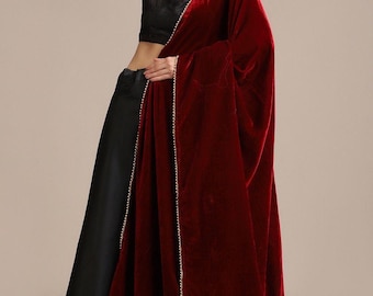 May include: A deep red velvet shawl with gold trim is draped over a black outfit. The shawl is the focal point, its rich color contrasting with the black clothing. The gold trim adds a touch of elegance to the design.