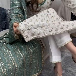 May include: An elegant outfit with a white and silver embellished top and matching trousers, with pearl detailing. A teal green sequined shawl adds a splash of colour. The outfit is paired with gold-coloured heels.