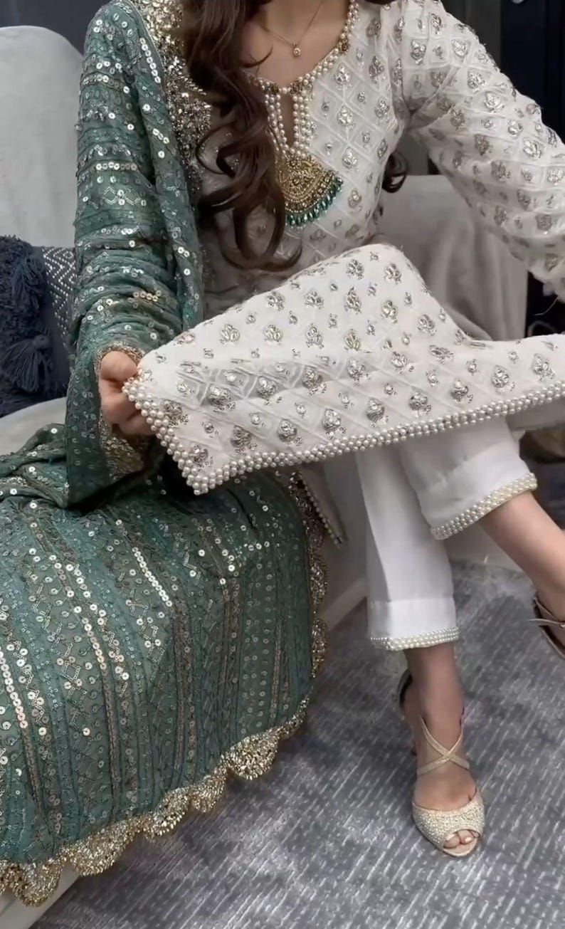 May include: An elegant outfit with a white and silver embellished top and matching trousers, with pearl detailing. A teal green sequined shawl adds a splash of colour. The outfit is paired with gold-coloured heels.