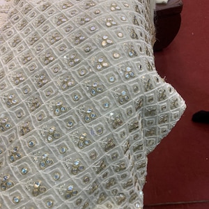 May include: Close-up of off-white fabric with a geometric pattern of squares and diamonds. The fabric is embellished with small, iridescent sequins and gold-coloured accents, creating a textured and decorative surface.