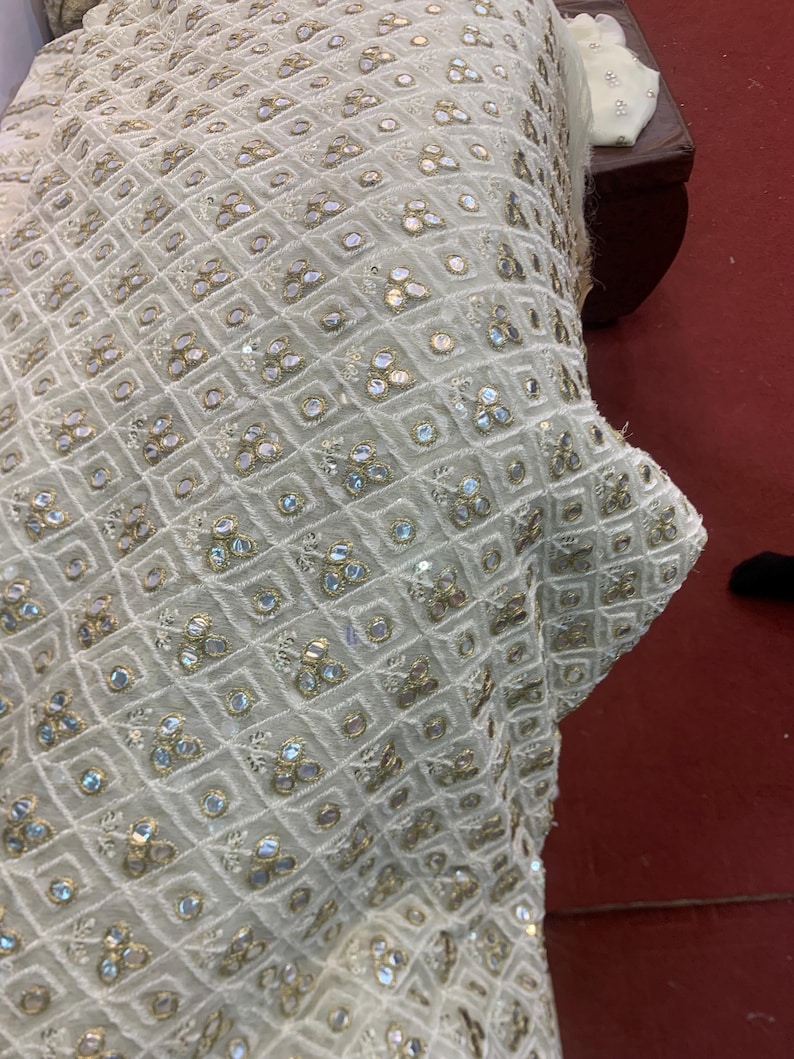 May include: Close-up of off-white fabric with a geometric pattern of squares and diamonds. The fabric is embellished with small, iridescent sequins and gold-coloured accents, creating a textured and decorative surface.