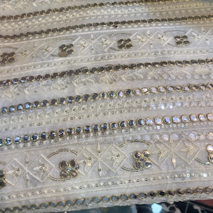 May include: Close-up of white fabric with detailed embroidery and embellishments. The design includes rows of mirrored accents, floral patterns, and geometric shapes, creating a textured and ornate appearance. The fabric has a delicate, luxurious feel.