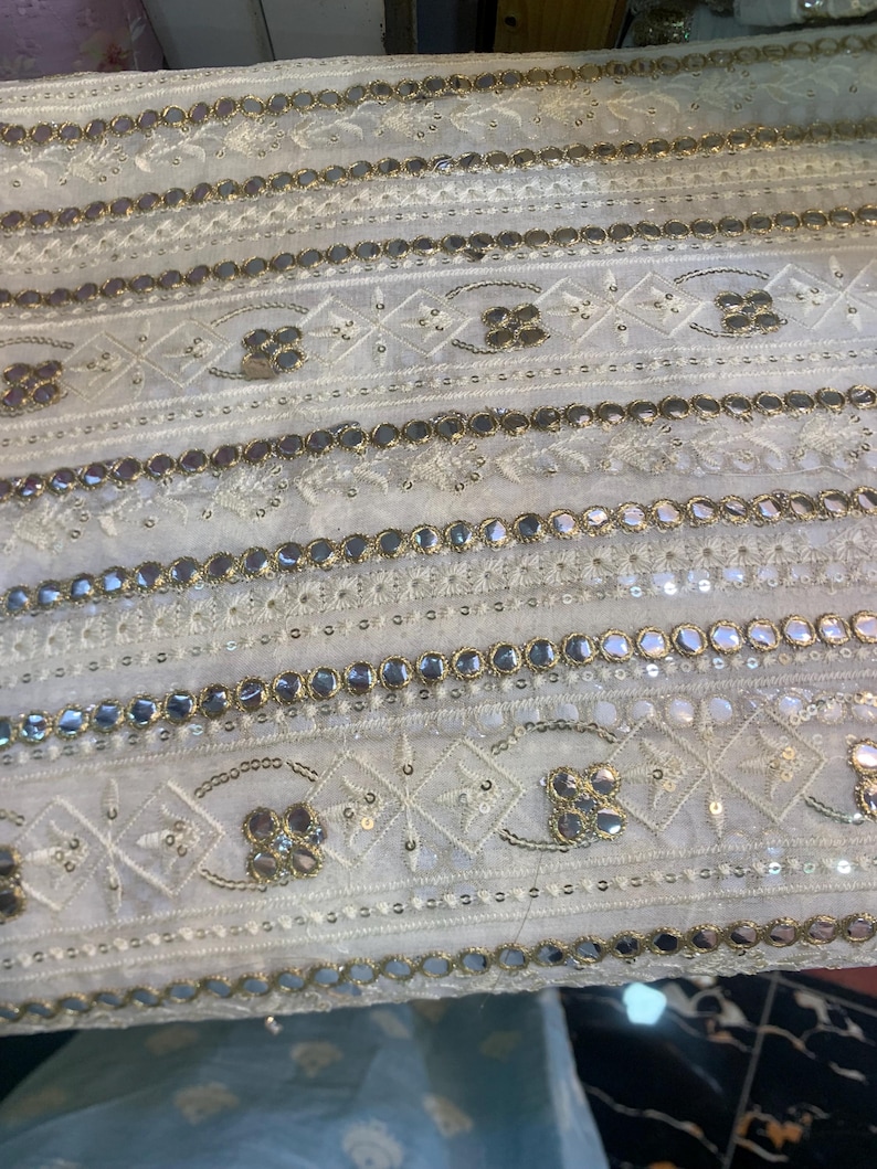 May include: Close-up of white fabric with detailed embroidery and embellishments. The design includes rows of mirrored accents, floral patterns, and geometric shapes, creating a textured and ornate appearance. The fabric has a delicate, luxurious feel.