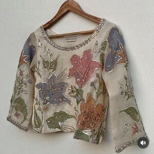 May include: Cream-colored long-sleeved blouse with floral embroidery in pink, orange, green, and blue. The blouse has a round neckline and is embellished with sequins. The brand name "MARIA BASHIR" is visible on the neckline.