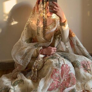 May include: An ornate, cream-colored traditional outfit with intricate embroidery and embellishments. The outfit features a sheer veil with gold trim and a matching purse. Pink and green floral patterns adorn the skirt, creating a luxurious and elegant aesthetic.