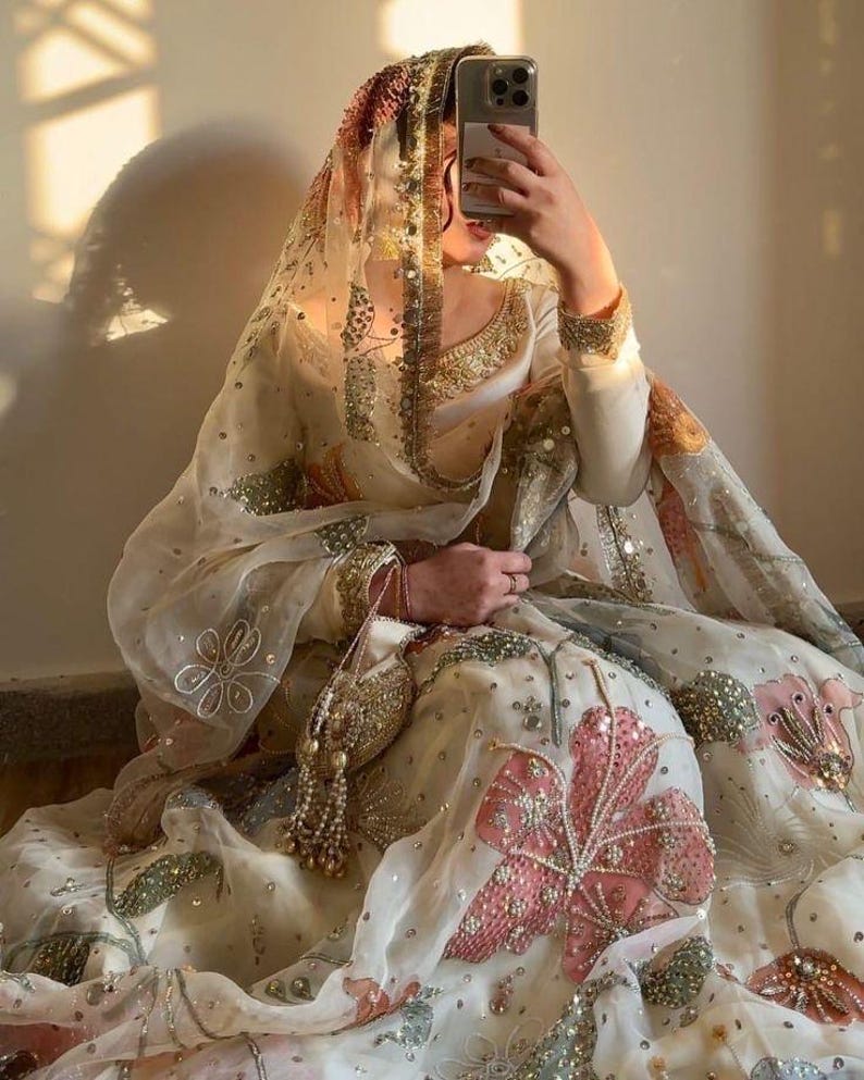 May include: An ornate, cream-colored traditional outfit with intricate embroidery and embellishments. The outfit features a sheer veil with gold trim and a matching purse. Pink and green floral patterns adorn the skirt, creating a luxurious and elegant aesthetic.