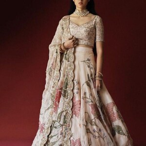 May include: An ivory-colored, floral-printed lehenga with a matching short-sleeved blouse and dupatta. The lehenga features a wide skirt with a silver border. The dupatta has a scalloped edge with floral and sequin detailing. The outfit is worn with jewelry.