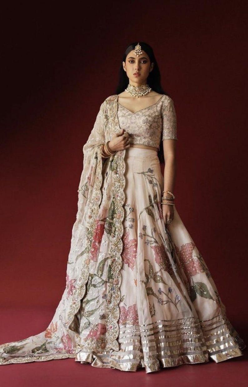 May include: An ivory-colored, floral-printed lehenga with a matching short-sleeved blouse and dupatta. The lehenga features a wide skirt with a silver border. The dupatta has a scalloped edge with floral and sequin detailing. The outfit is worn with jewelry.