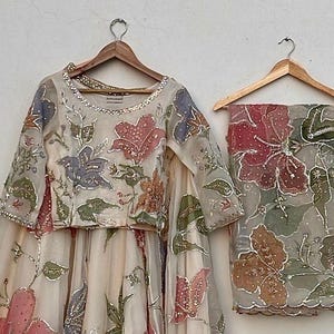 May include: A cream-colored, floral-embroidered outfit, including a long-sleeved top and a skirt, hangs on a wooden hanger. The outfit features pink, blue, and green floral designs with silver accents. A matching scarf with similar floral embroidery hangs on a separate hanger.