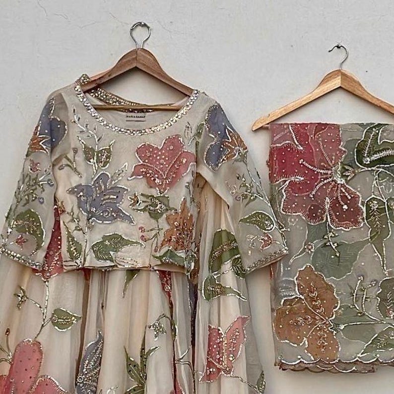 May include: A cream-colored, floral-embroidered outfit, including a long-sleeved top and a skirt, hangs on a wooden hanger. The outfit features pink, blue, and green floral designs with silver accents. A matching scarf with similar floral embroidery hangs on a separate hanger.