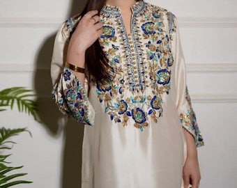 May include: Cream-colored, long-sleeved tunic with intricate floral embroidery in blue, gold, and teal. The embroidery adorns the neckline, cuffs, and bodice. The tunic has a mandarin collar and a relaxed fit, suitable for special occasions.