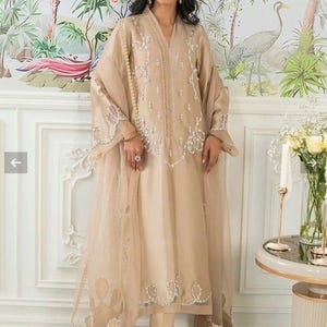 Puede incluir: Un vestido formal beige bordado con un chal transparente a juego. El vestido presenta intrincados adornos plateados a lo largo del escote, las mangas y el dobladillo. El conjunto se combina con pantalones y tacones beige a juego. El fondo presenta un mural floral.