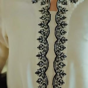 以下が含まれることがあります： ネックラインと中央に複雑な黒い花の刺繍が施されたクリーム色のチュニック。長袖の衣服は、正方形のネックラインと、フォーマルまたはお祝いの機会に適した、詳細で対称的なデザインが特徴です。