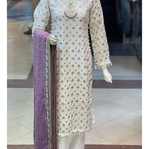May include: White embroidered tunic and matching trousers with a purple scarf. The tunic features a diamond pattern with gold accents, long sleeves, and beaded trim. The scarf is a solid purple with gold trim.