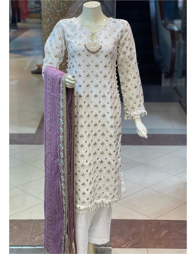 May include: White embroidered tunic and matching trousers with a purple scarf. The tunic features a diamond pattern with gold accents, long sleeves, and beaded trim. The scarf is a solid purple with gold trim.