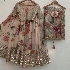 May include: A blush-colored, floral-printed ensemble featuring a blouse and skirt with gold trim. The outfit includes a matching scarf and small purse, all adorned with intricate floral patterns. The blouse has a V-neck and short sleeves.