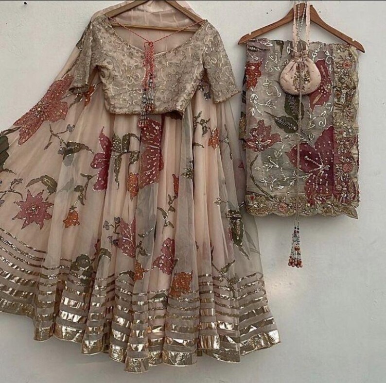 May include: A blush-colored, floral-printed ensemble featuring a blouse and skirt with gold trim. The outfit includes a matching scarf and small purse, all adorned with intricate floral patterns. The blouse has a V-neck and short sleeves.