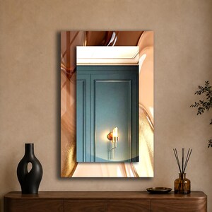 May include: A rectangular wall mirror featuring a central clear glass panel surrounded by a decorative border with abstract, flowing brown, gold, and cream-colored patterns. The mirror reflects a teal paneled wall and a lit wall sconce.