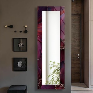 May include: A tall, rectangular mirror with a floral design in shades of burgundy and purple. The mirror has a white reflective surface and is mounted on a gray wall. A small plant is visible at the bottom of the mirror.