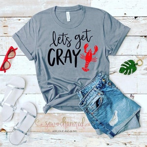 May include: Gray t-shirt with the text "Let's get Cray" and a red crawfish illustration. The shirt is on a wooden surface with a pair of white sandals, a pair of denim shorts, and a green leaf.