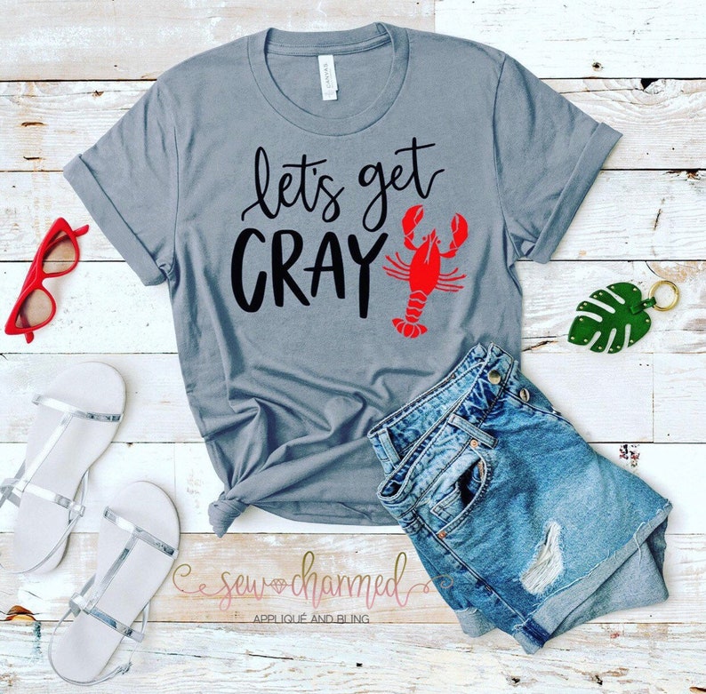 May include: Gray t-shirt with the text "Let's get Cray" and a red crawfish illustration. The shirt is on a wooden surface with a pair of white sandals, a pair of denim shorts, and a green leaf.