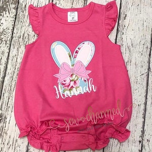 May include: Pink baby onesie with a floral bunny applique and the name "Hannah" embroidered on the front. The onesie has ruffle sleeves and a ruffle bottom.