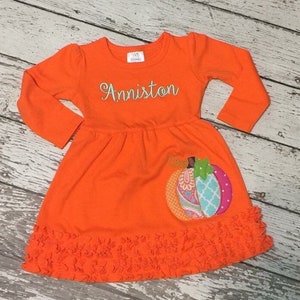 May include: Orange long-sleeved dress with a ruffled hem and a colorful pumpkin applique. The dress is personalized with the name "Anniston".