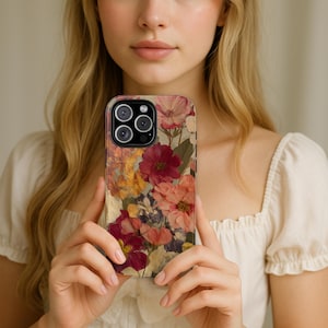 Puede incluir: Funda de teléfono inteligente con un collage de flores secas en tonos rojos, rosas, amarillos y morados. La funda tiene un fondo transparente, lo que permite ver las lentes de la cámara del teléfono. El diseño floral está prensado y dispuesto.