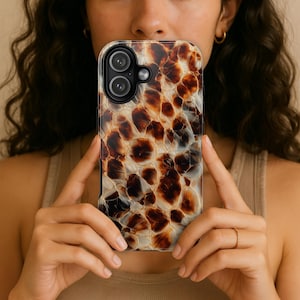 Op de afbeelding: Een smartphonehoesje met een bruin en wit schildpadpatroon. De hoes heeft een glanzende afwerking en een camera-uitsparing. De hoes wordt voor het gezicht van een persoon gehouden.